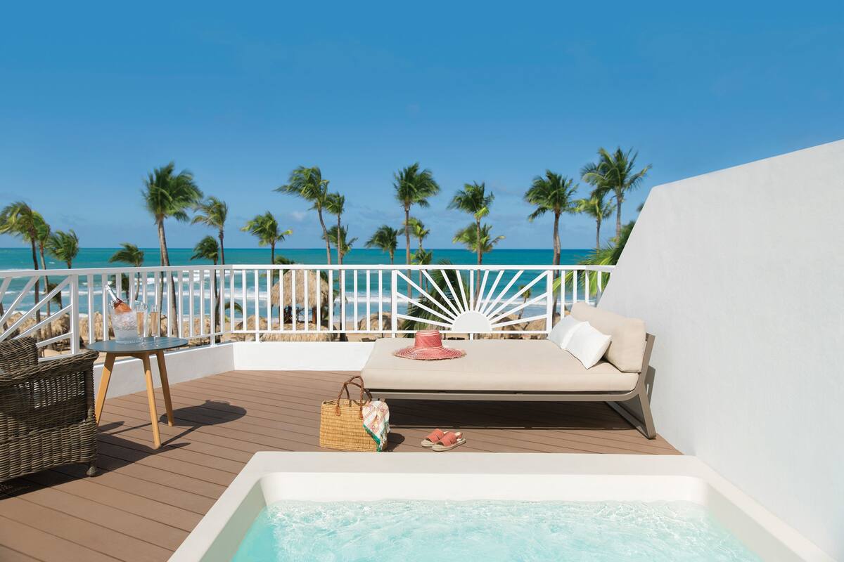Une terrasse avec un canapé, une table et un jacuzzi, donnant sur une plage bordée de palmiers.