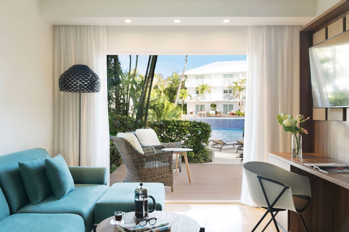 Une chambre d’hôtel moderne, équipée d’un canapé, d’une table basse et d’un bureau avec une chaise. Une grande fenêtre offre une vue sur une piscine et un immeuble.