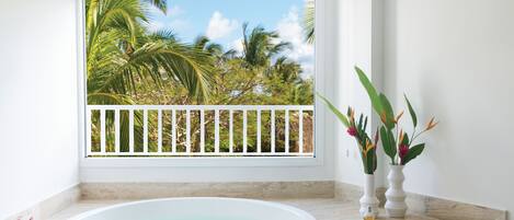A bathroom with a large bathtub, a window with a view of palm trees, and two vases with flowers.