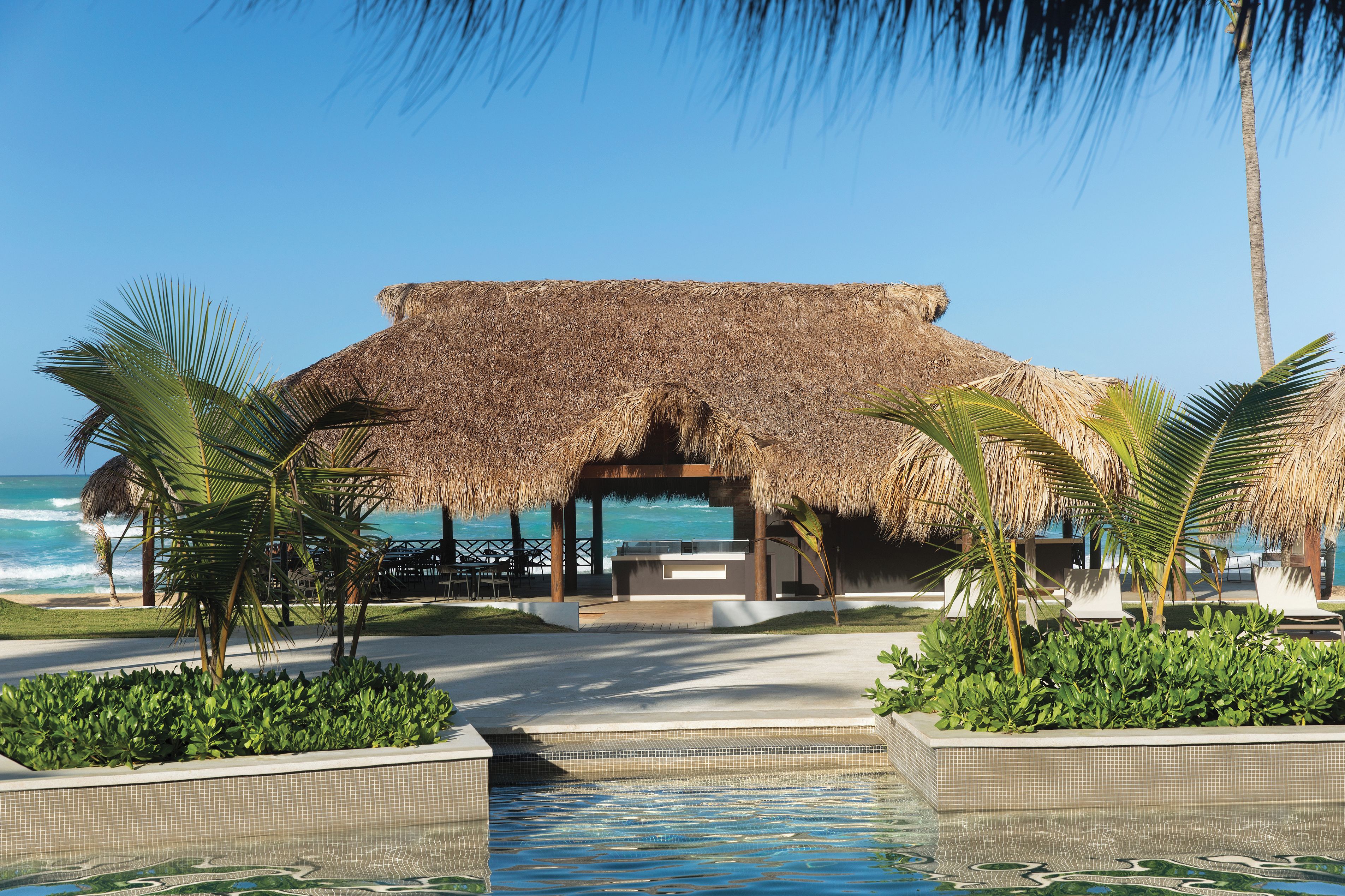 A pool area with a thatched-roof structure, palm trees, and a view of the ocean.