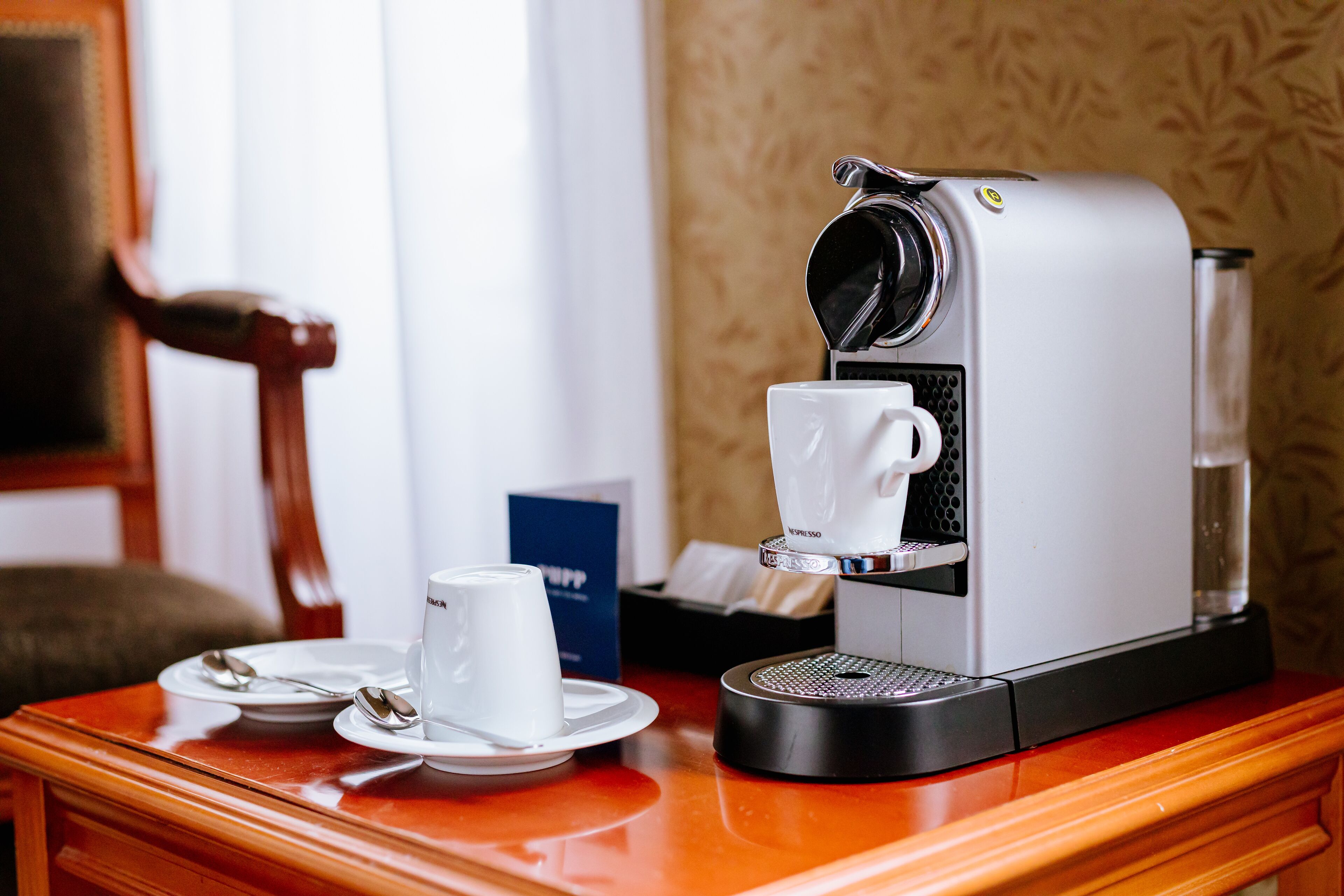 Une machine à café, une tasse avec sa soucoupe, des cuillères et une boîte, le tout posé sur une table en bois.