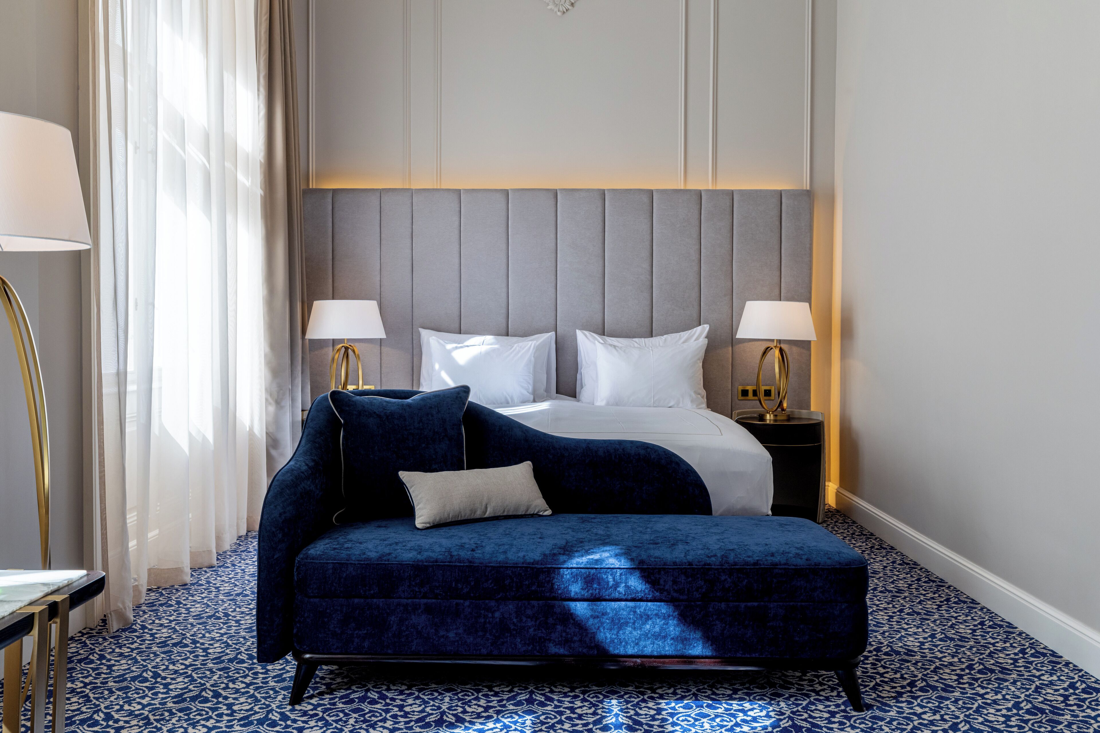 Une chambre d’hôtel avec un canapé bleu recouvert de tissu, un lit avec du linge de maison blanc, deux lampes de chevet et une moquette à motifs.