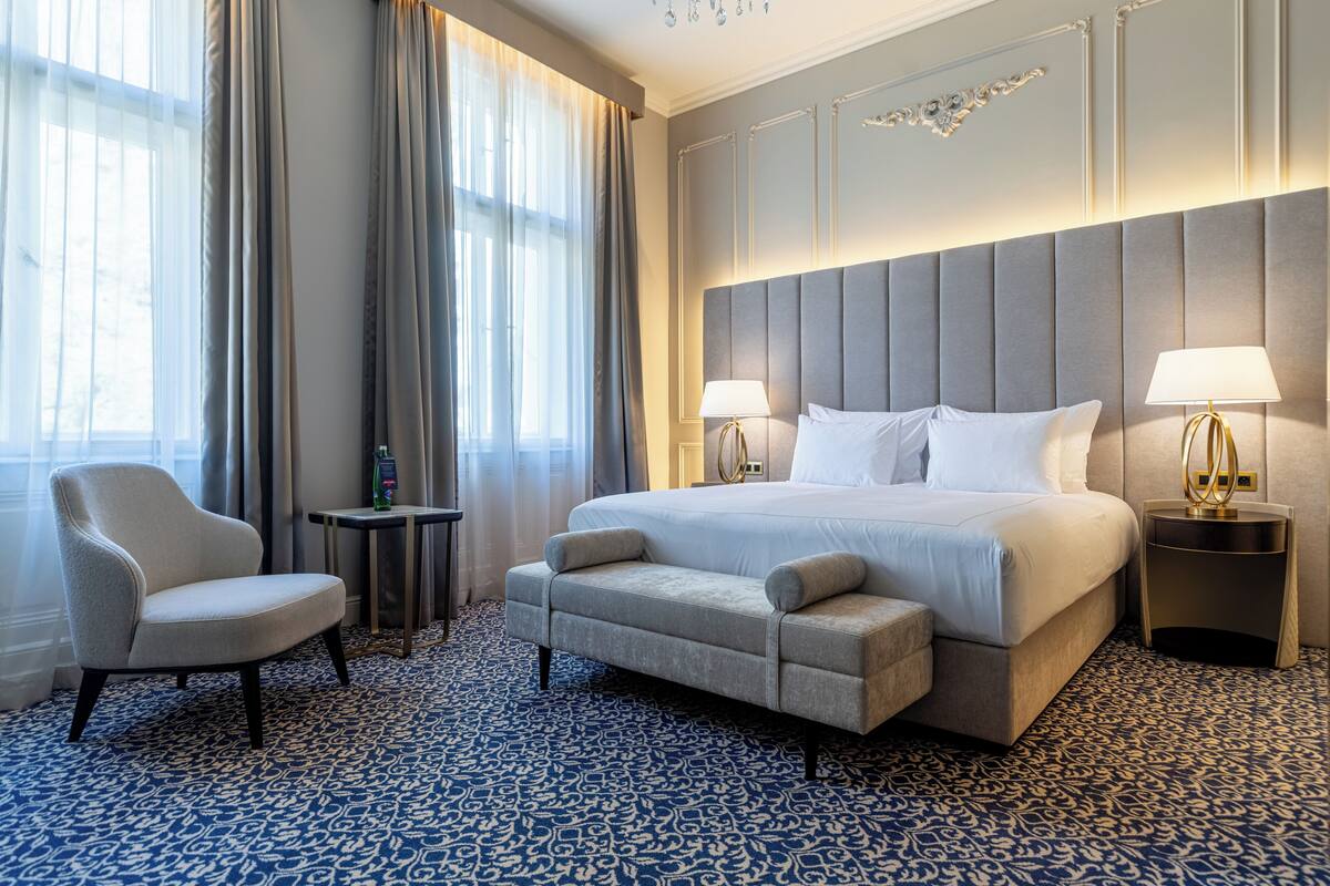 Quarto de hotel com uma cama grande, duas mesas de cabeceira, uma cadeira e uma mesinha.