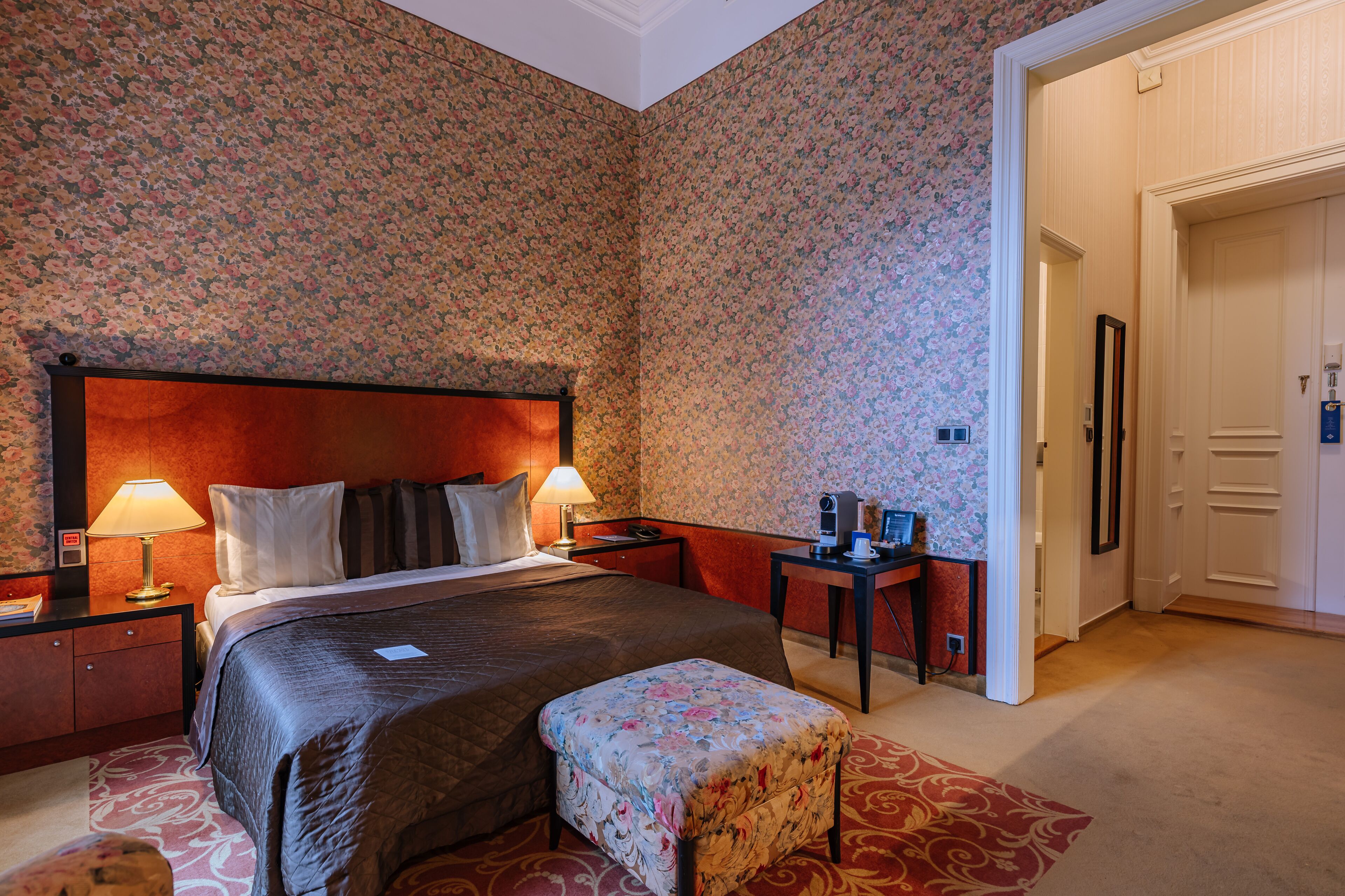 A hotel room with a large bed, two bedside lamps, a desk with a computer, and a small table with a lamp.