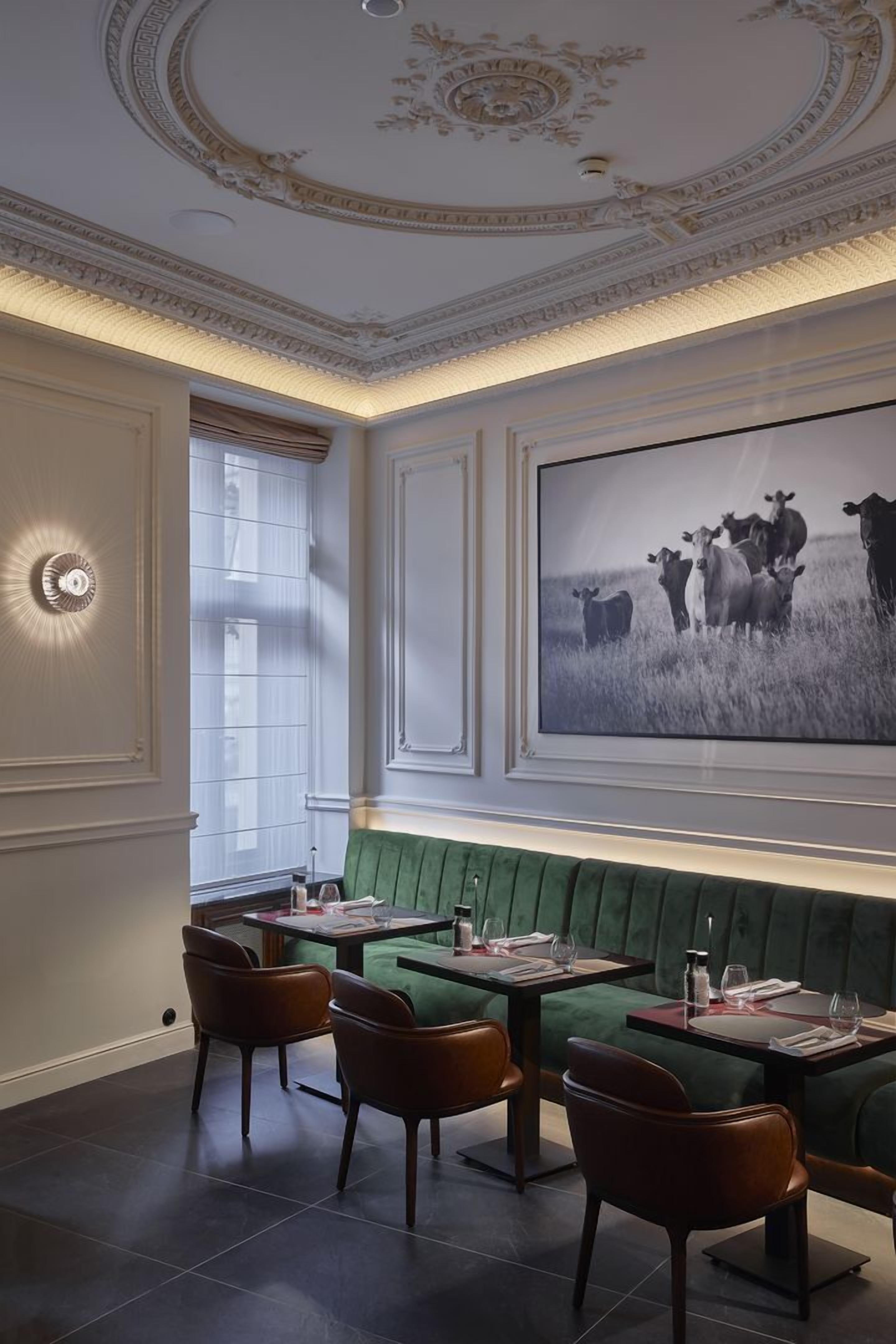 A dining area with a green upholstered bench, wooden chairs, and a large framed photograph of cows on the wall.