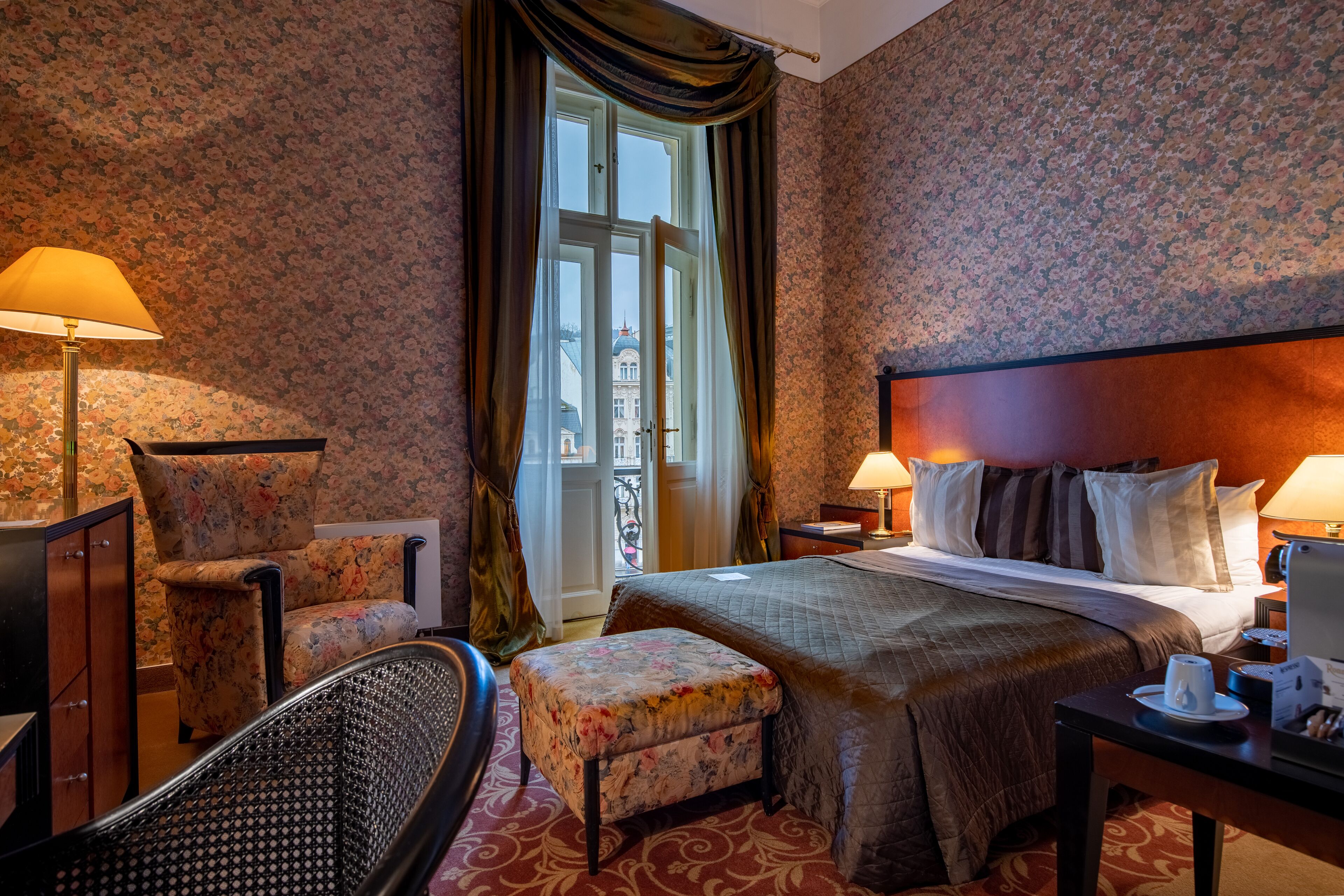 Une chambre d’hôtel avec un lit, deux lampes, une chaise, un pouf, un bureau et une vue sur un immeuble par la fenêtre.