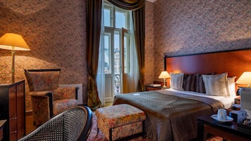 Une chambre d’hôtel avec un lit, deux lampes, une chaise, un pouf, un bureau et une vue sur un immeuble par la fenêtre.