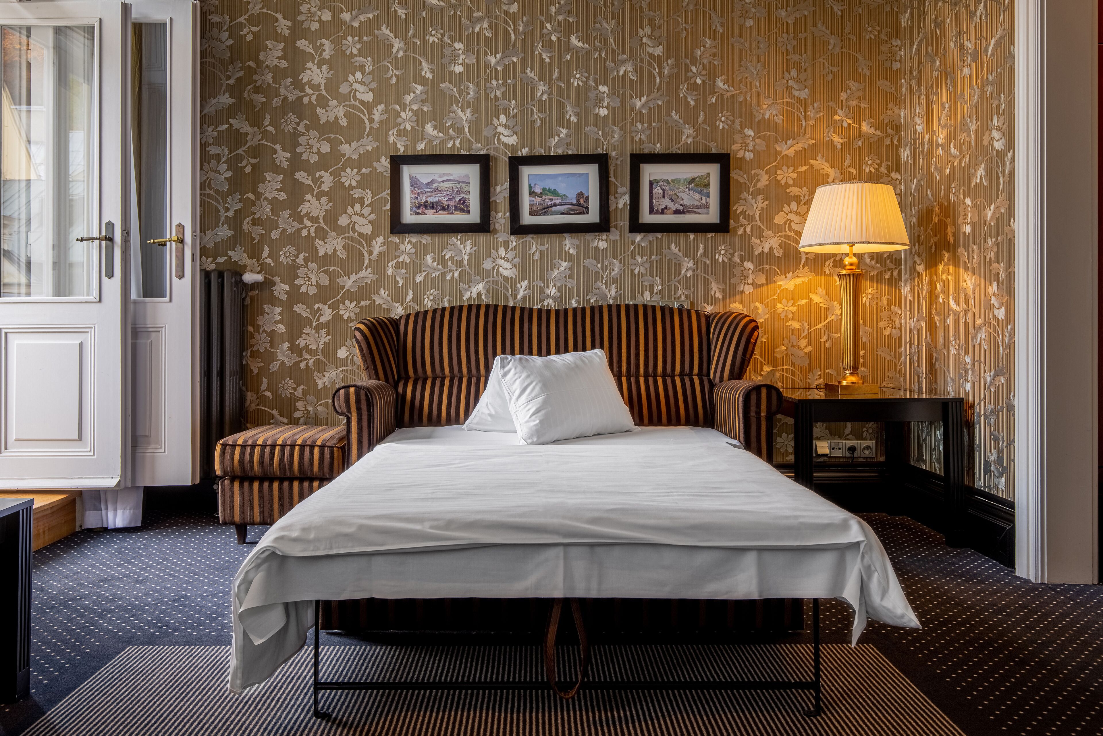 Une chambre d’hôtel avec un lit, un fauteuil à rayures, une petite table avec une lampe et trois tableaux encadrés accrochés au mur.