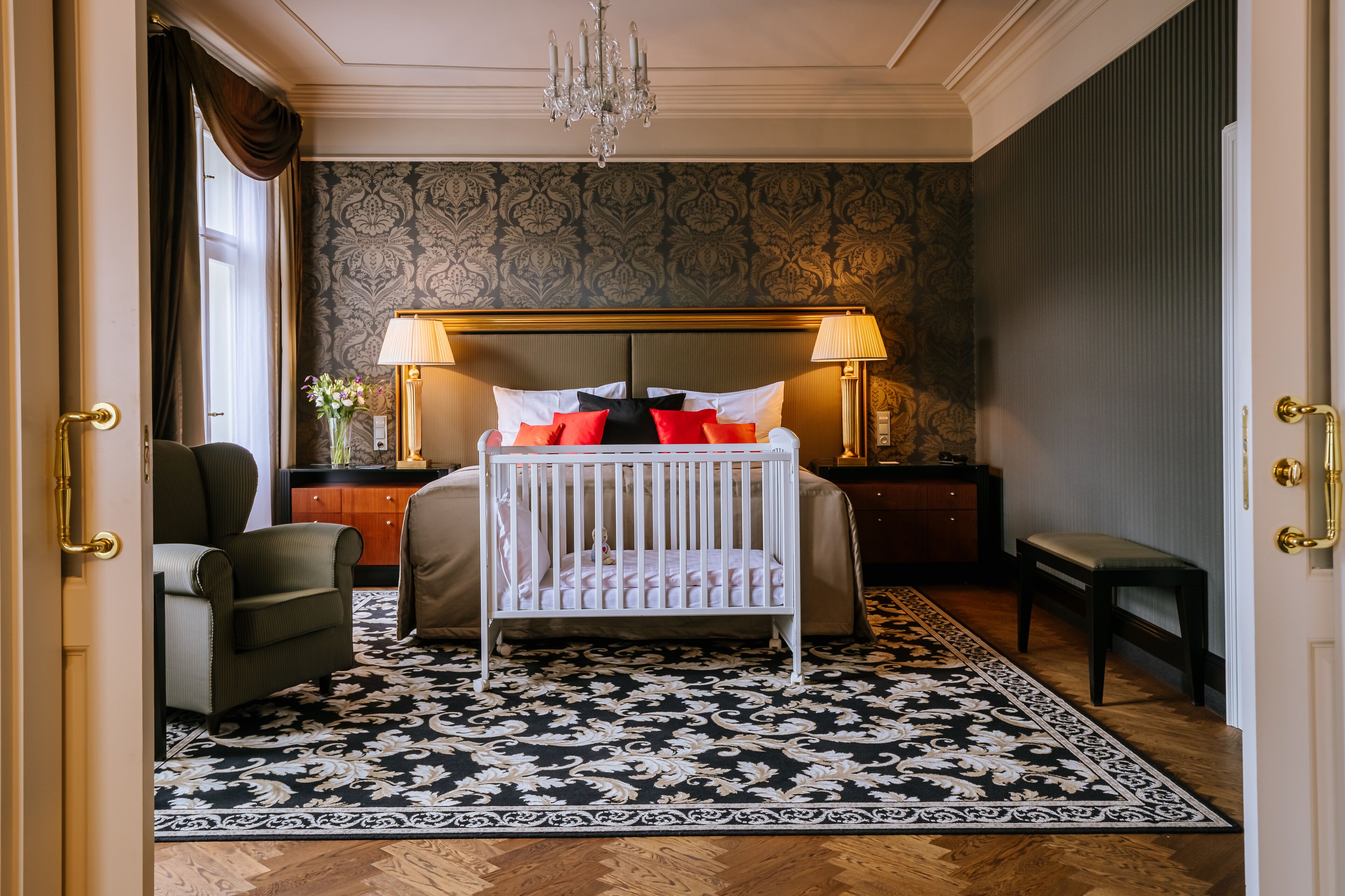 A bedroom with a crib, a bedside table with a lamp, a chair, and a bench.