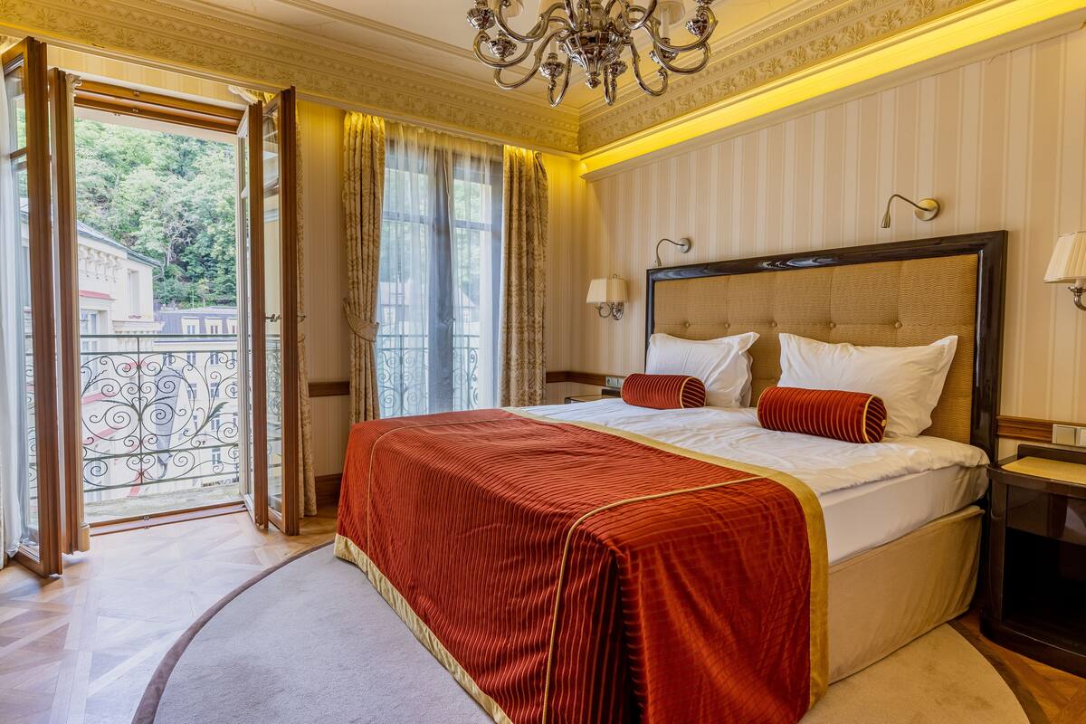 Quarto de hotel com uma cama grande, um candelabro, uma varanda com grade de ferro e vista para o verde lá fora.