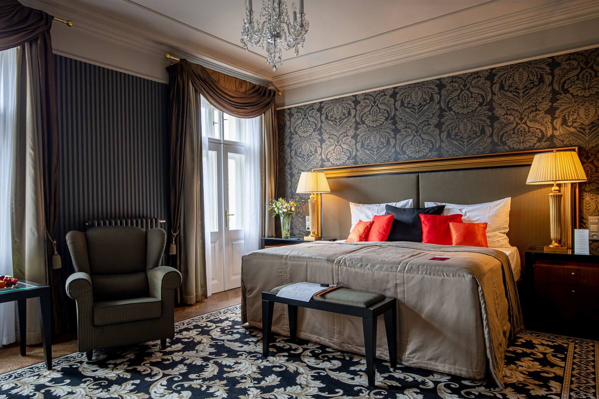 Quarto de hotel com uma cama grande, uma cadeira, uma pequena mesa e um candelabro.