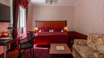 Ein Hotelzimmer mit Bett, Schreibtisch, Sessel, Sofa, einer Kandelaber und Wandtapete mit Muster.