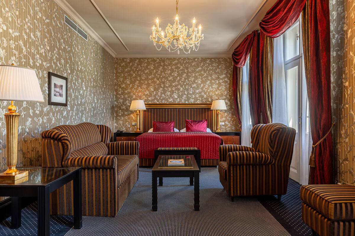 Quarto de hotel com uma cama grande, duas poltronas, uma mesa pequena e um candelabro.
