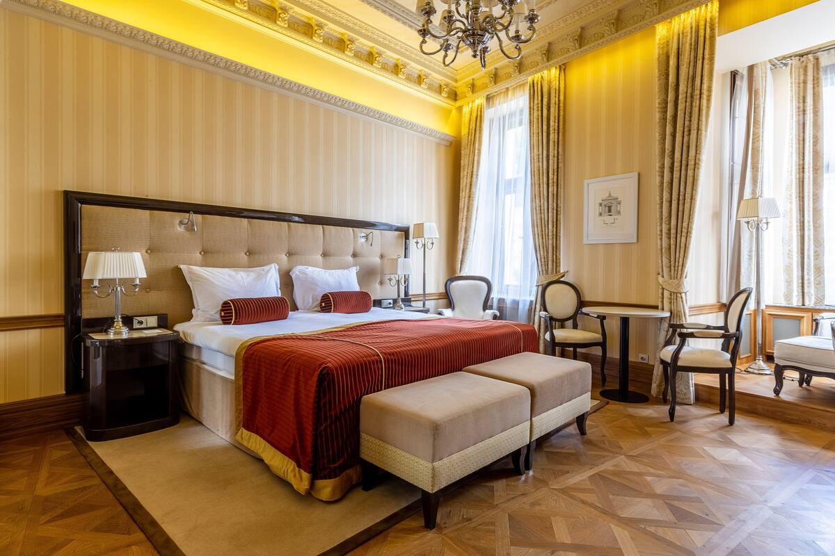 Quarto de hotel espaçoso com uma cama grande, área de estar com mesa e cadeiras, e vista da cidade por grandes janelas.