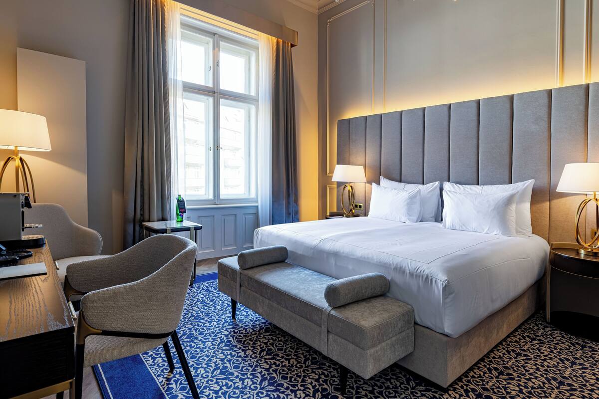Quarto de hotel com uma cama grande, duas mesas de cabeceira com luminárias, uma escrivaninha com cadeira e um tapete estampado.