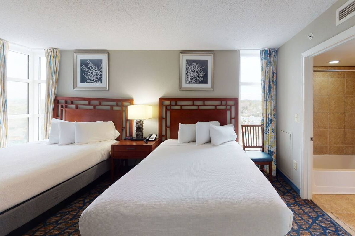 A hotel room with two beds, a nightstand with a lamp, a framed picture on the wall, and a shower area.