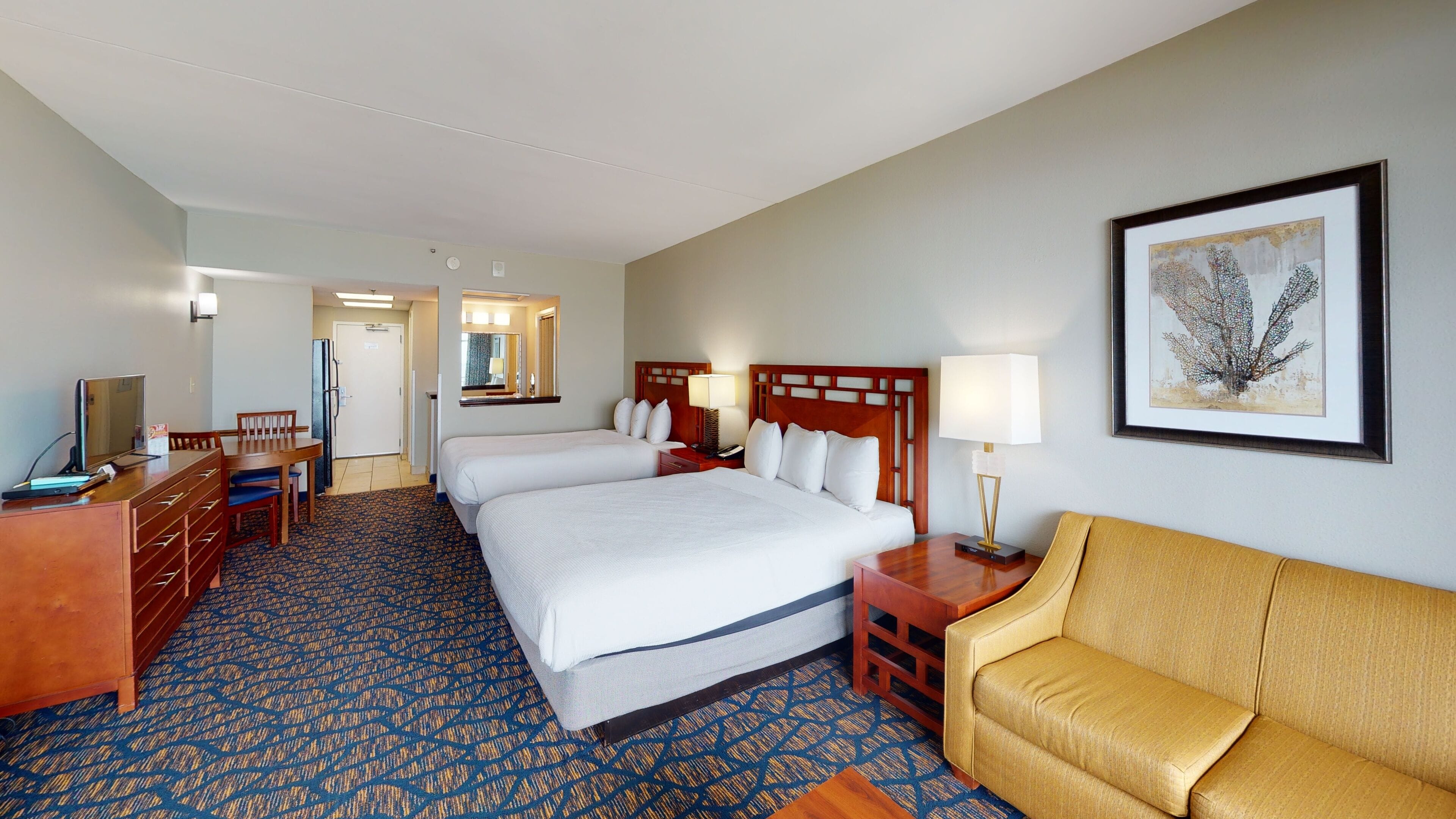 A hotel room with two beds, a desk with a computer, a chair, a sofa, and a framed artwork on the wall.