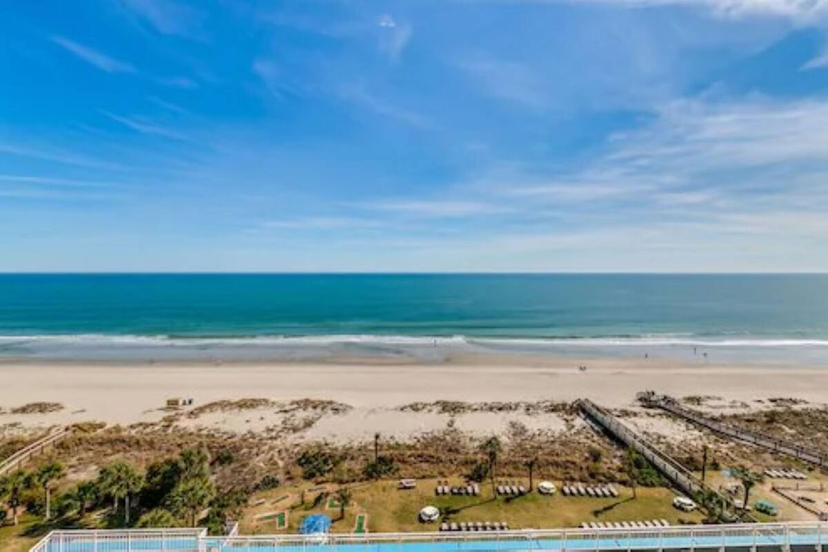 A beach view with a clear sky, a sandy beach, and a pool area with lounge chairs.