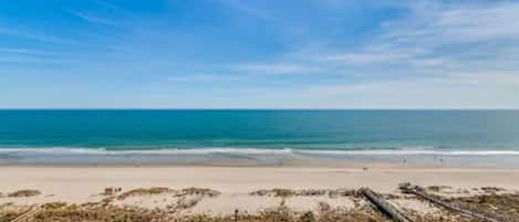 Blick auf einen Strand mit klarem Himmel, feinem Sand und einem Poolbereich mit Liegestühlen.