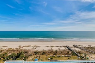 Vista de una playa con cielo despejado, arena dorada y un área de piscina con tumbonas.