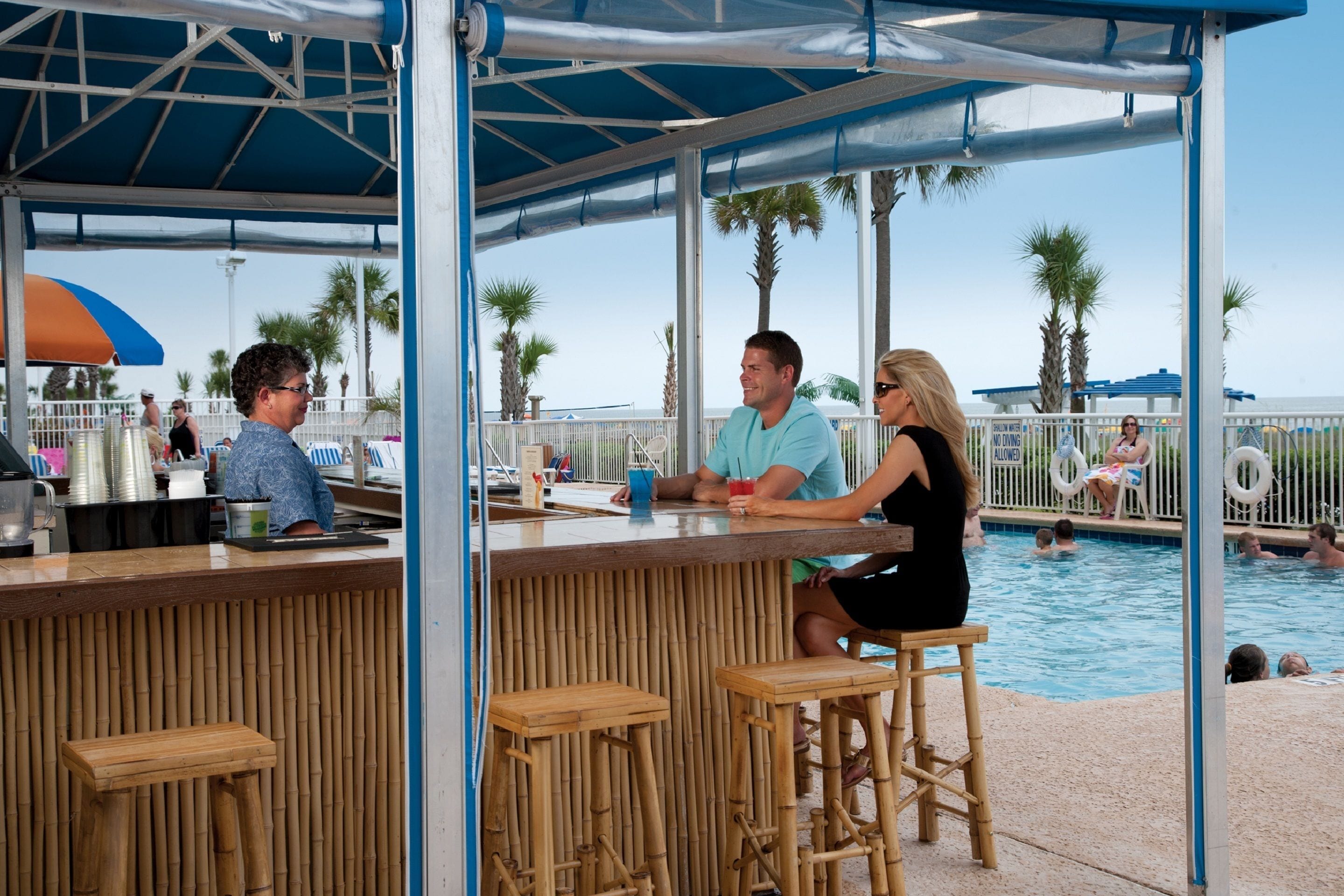 Un espace bar au bord de la piscine, avec des tabourets en bambou, un comptoir et une piscine en arrière-plan.