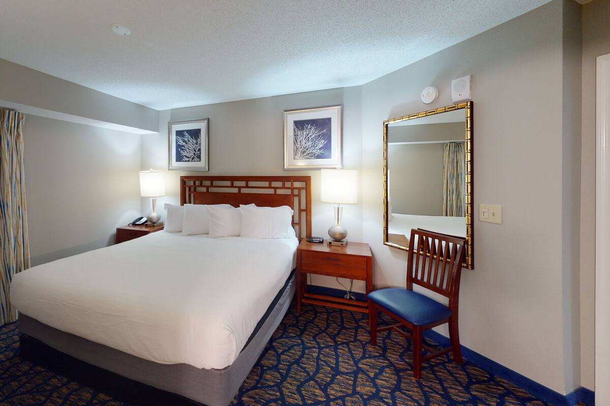 A hotel room with a large bed, two bedside tables with lamps, a chair, a mirror, and artwork on the walls.