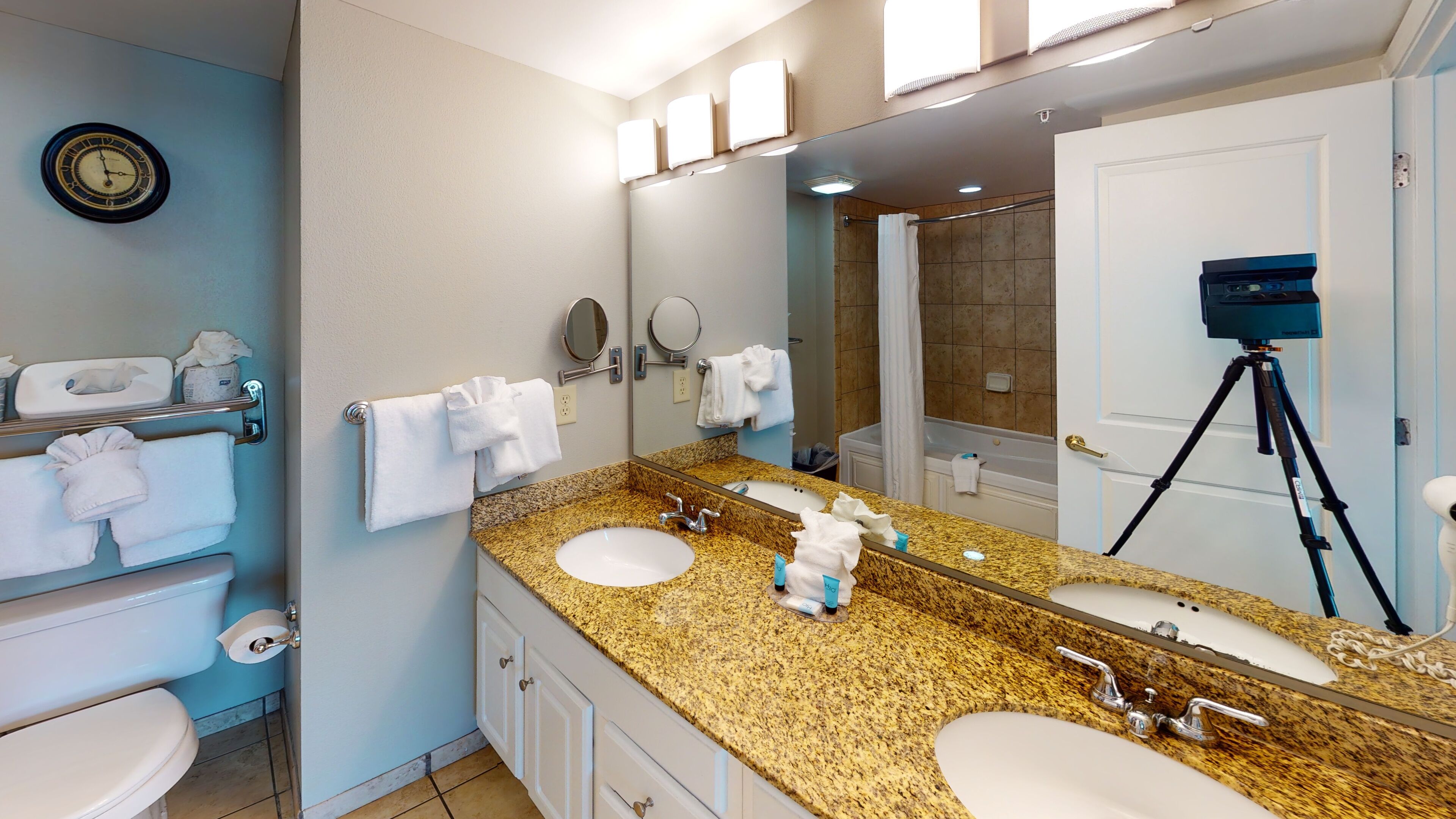 A hotel bathroom with two sinks, a large mirror, a clock, and a camera on a tripod.