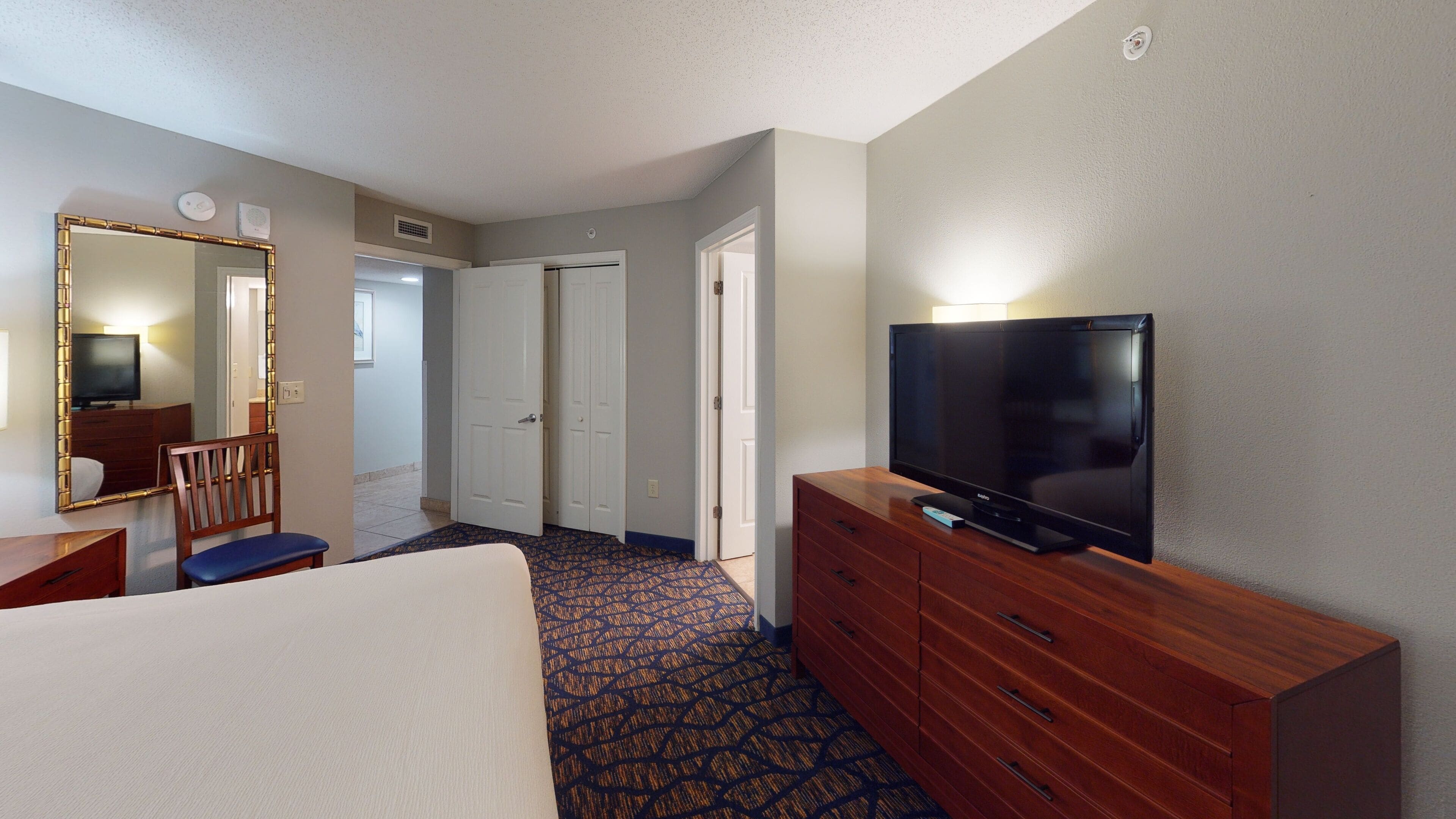 A hotel room with a bed, a wooden dresser, a television, a mirror, and a patterned carpet.