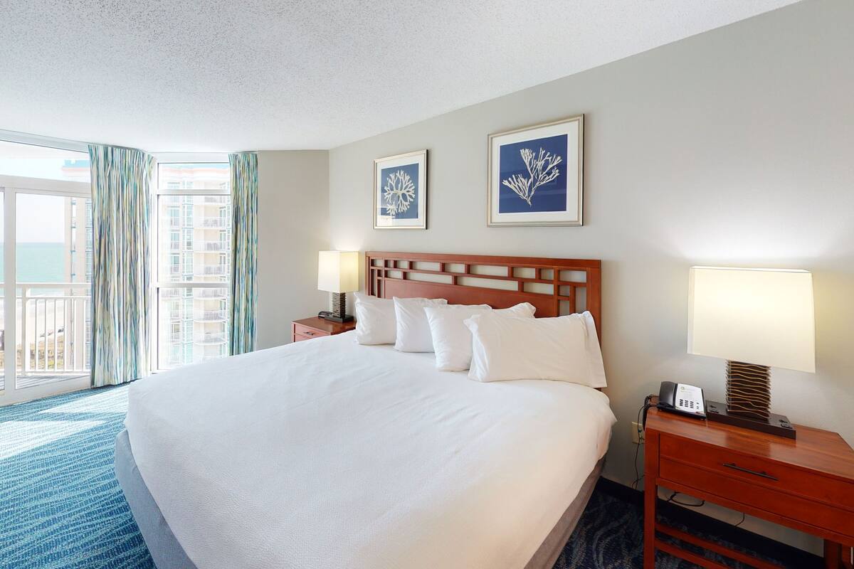 A hotel room with a large bed, two bedside tables, a chair, a desk, and a balcony with curtains.