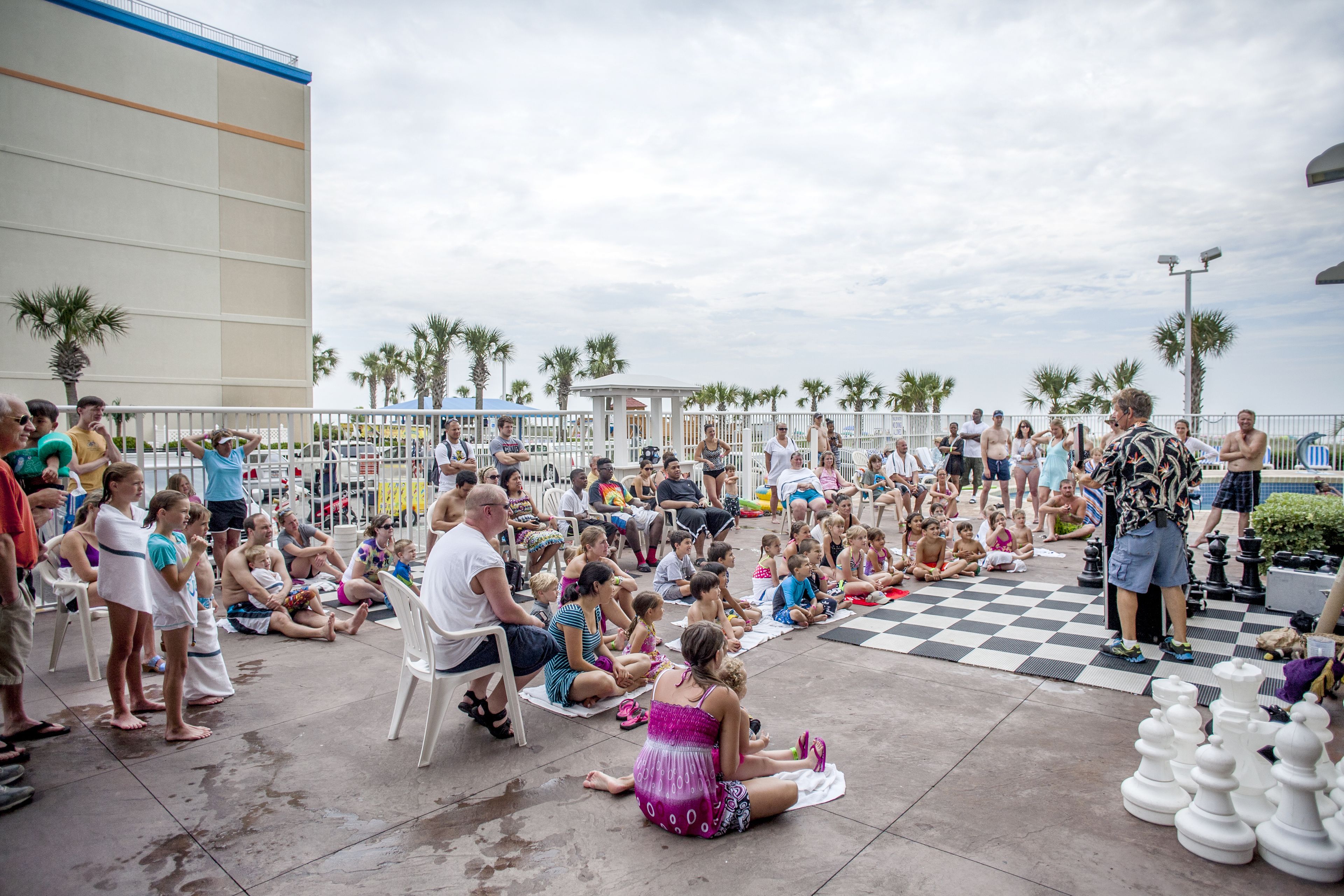 A group of people sitting on the ground outdoors, some on chairs, with a checkered floor and palm trees in the background.