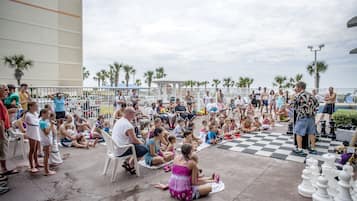 A group of people sitting on the ground outdoors, some on chairs, with a checkered floor and palm trees in the background.