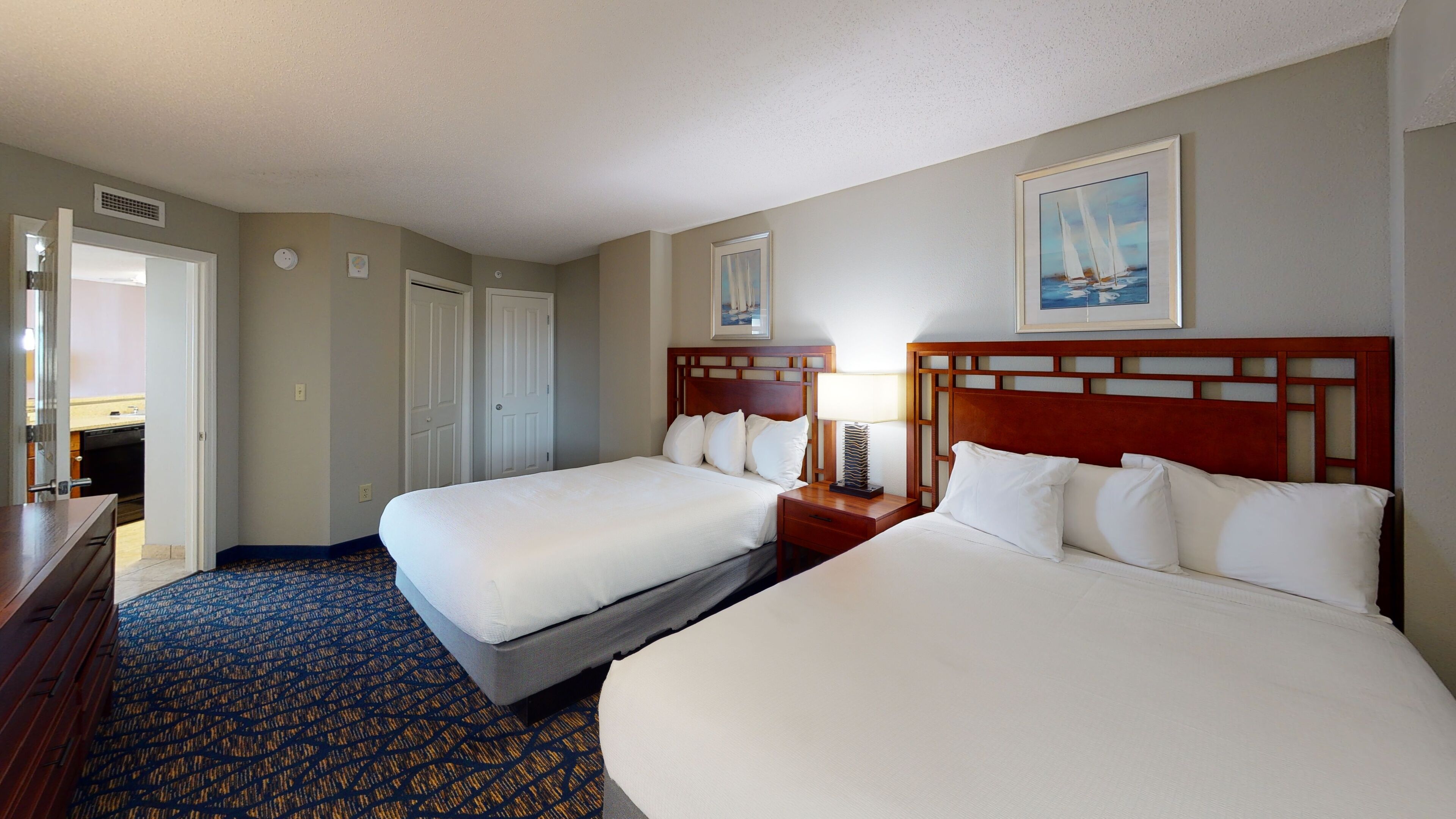 A hotel room with two beds, a wooden headboard, a bedside table with a lamp, a framed picture on the wall, and a patterned carpet.