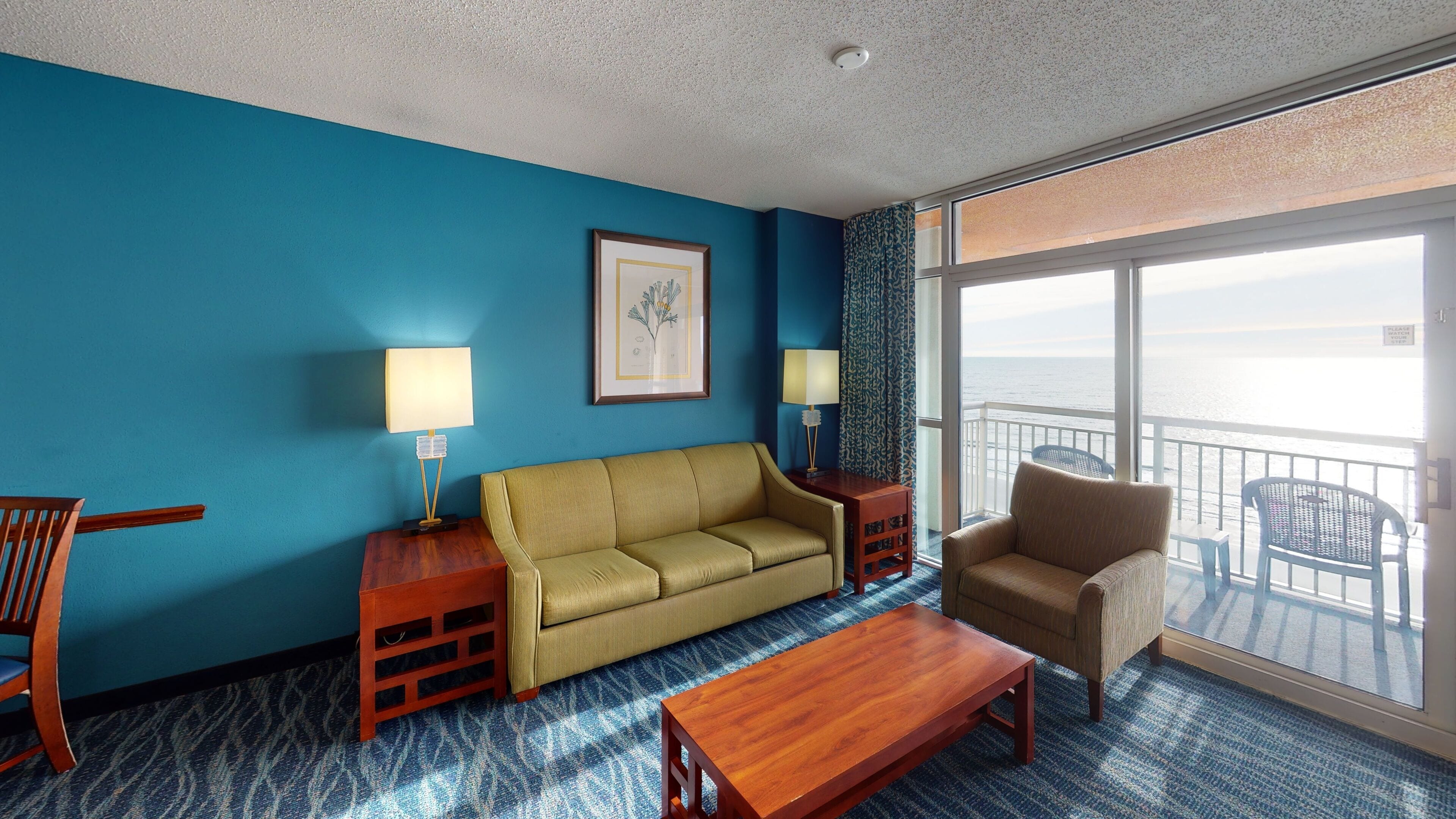 Habitación de hotel con pared azul, un sofá, una silla, una mesa de centro de madera y un balcón con vistas al mar.