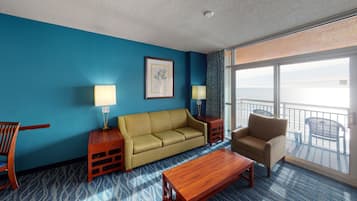 Habitación de hotel con pared azul, un sofá, una silla, una mesa de centro de madera y un balcón con vistas al mar.