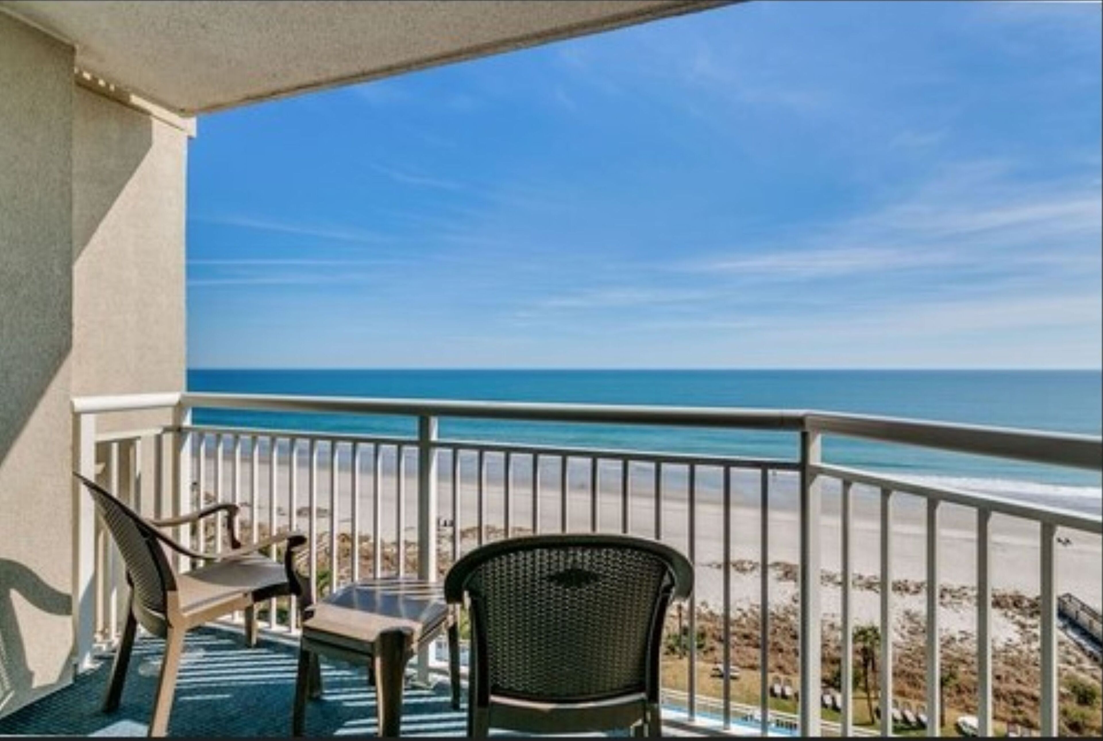 A balcony with two chairs and a table, overlooking a beach with a clear sky.