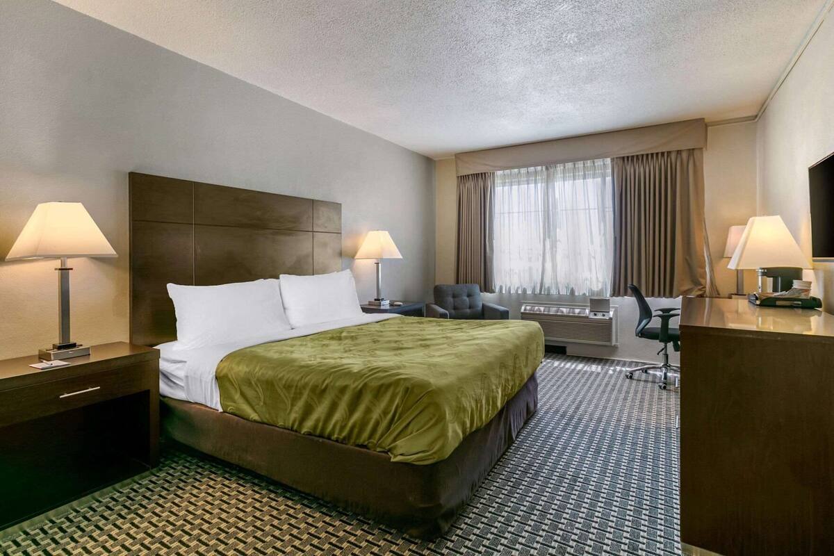 Habitación de hotel con cama, dos mesitas de noche con lámparas, escritorio con silla, ventana con cortinas y televisor montado en la pared.