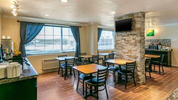 A restaurant with multiple tables and chairs, a stone pillar, a flat-screen TV mounted on the wall, and a view of the outside through large windows.