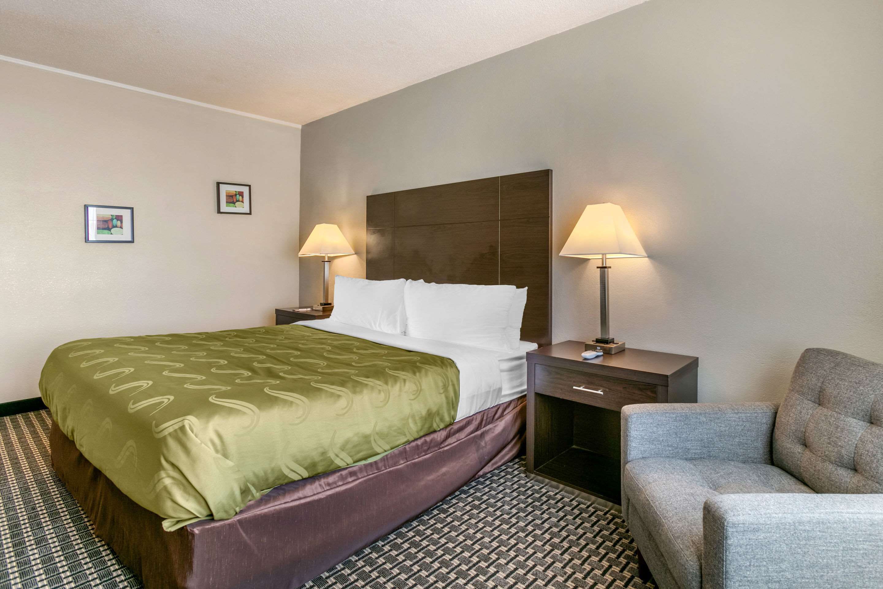 Hotellihuone, jossa on suuri sänky, kaksi yöpöytälamppua, yöpöytä, tuoli ja kuviollinen matto.
