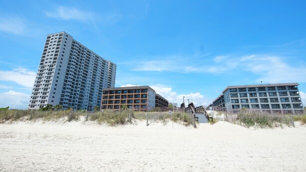 Ett hotell vid stranden med flera höga byggnader, en sandstrand i förgrunden och en klarblå himmel.