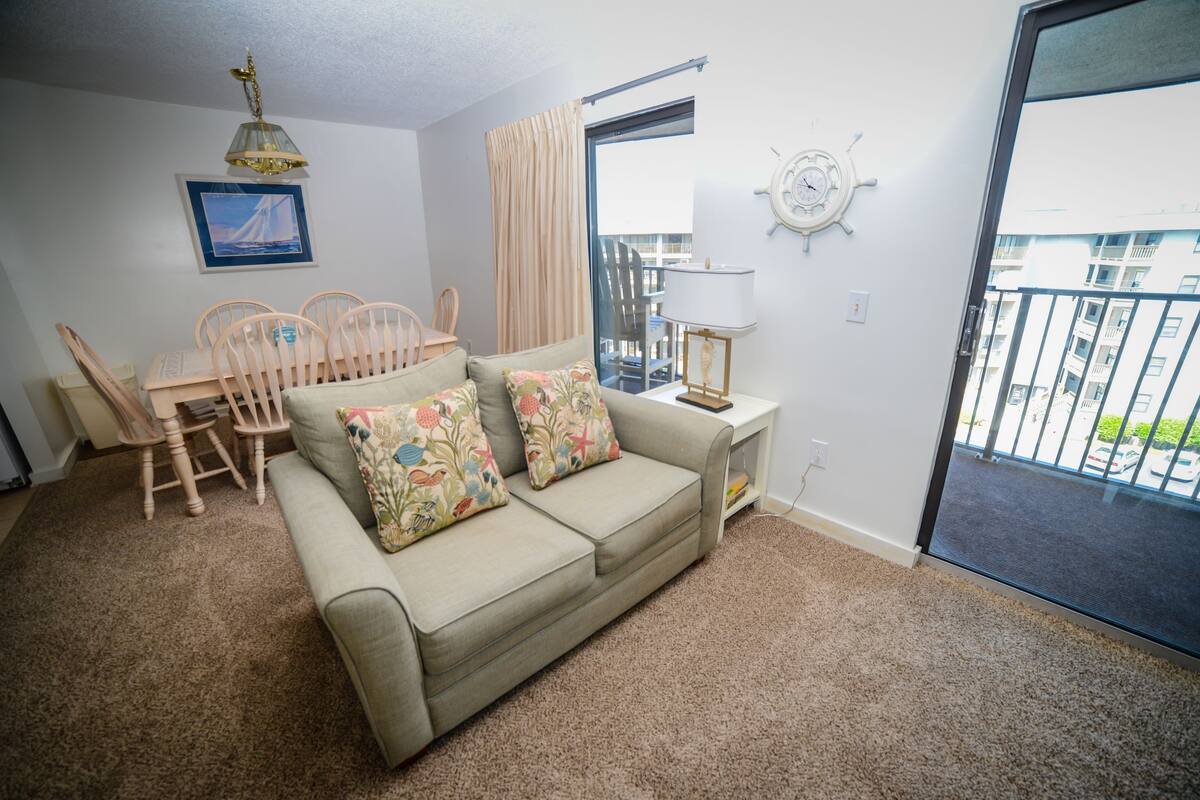A living room with a beige sofa, floral cushions, a wooden dining table with chairs, a lamp, a wall clock, and a view of the outside balcony.