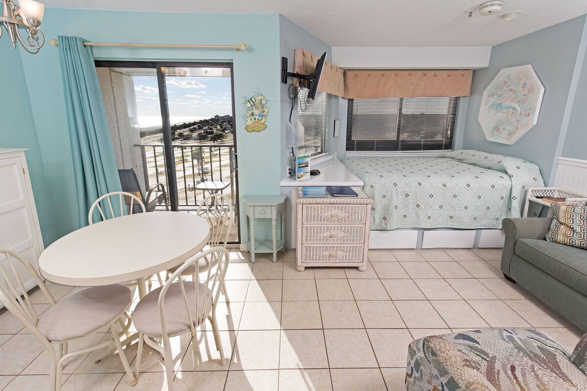A room with a dining table, chairs, a bed, a sofa, and a view through a sliding door.