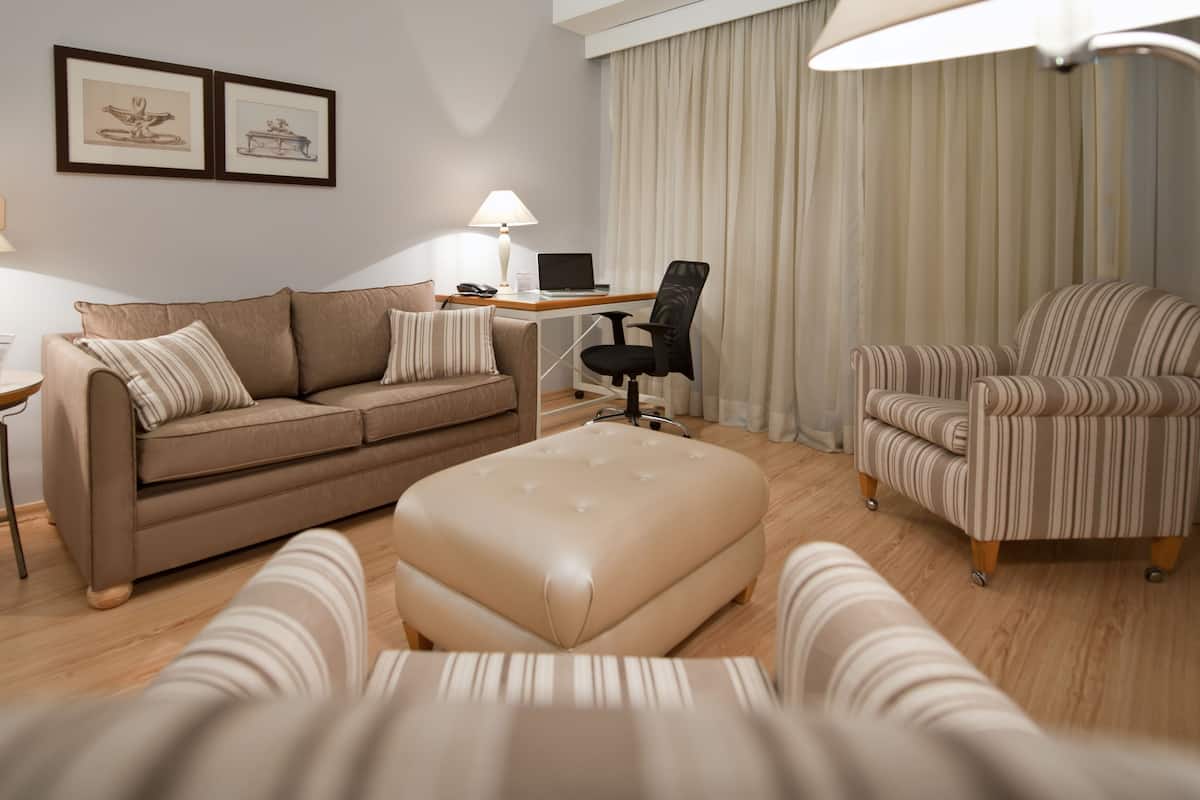 Ein modernes Hotelzimmer mit Sofa, Ohrensessel, Hocker, Schreibtisch und Lampe.