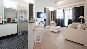 A modern hotel room with a kitchenette, a living area with a sofa and armchair, a flat-screen TV, and a large window with curtains. - HB Hotels Sequóia Alphaville (Barueri)