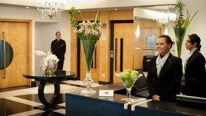 A reception area with a reception desk, two vases with flowers, a chandelier, and a man in uniform in the background. - HB Hotels Sequóia Alphaville (Barueri)