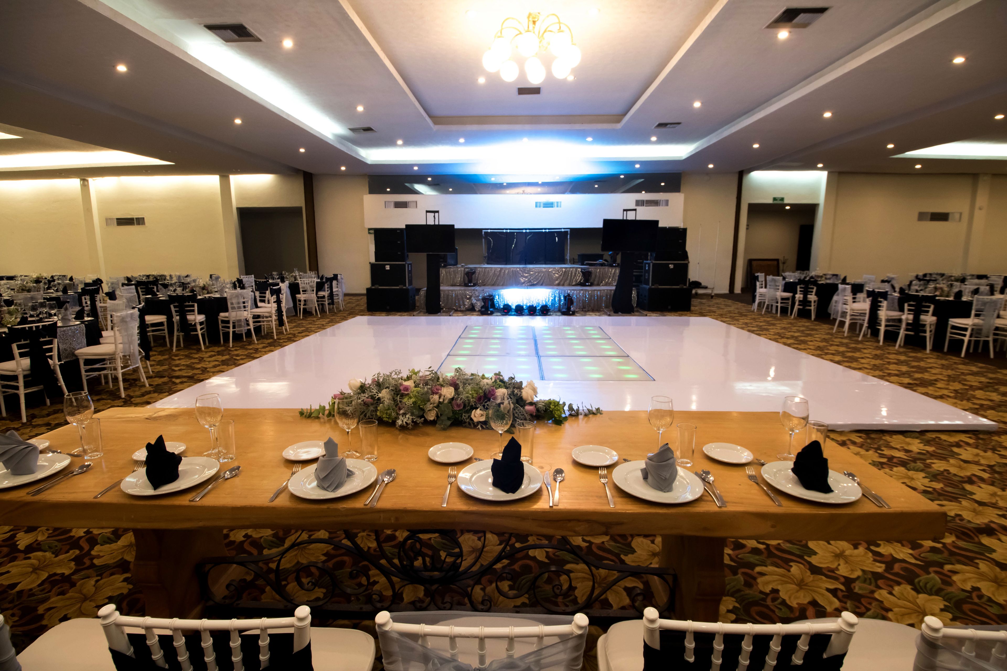 Un salón de banquetes con mesas dispuestas para un evento formal, un escenario con oradores y un suelo alfombrado.