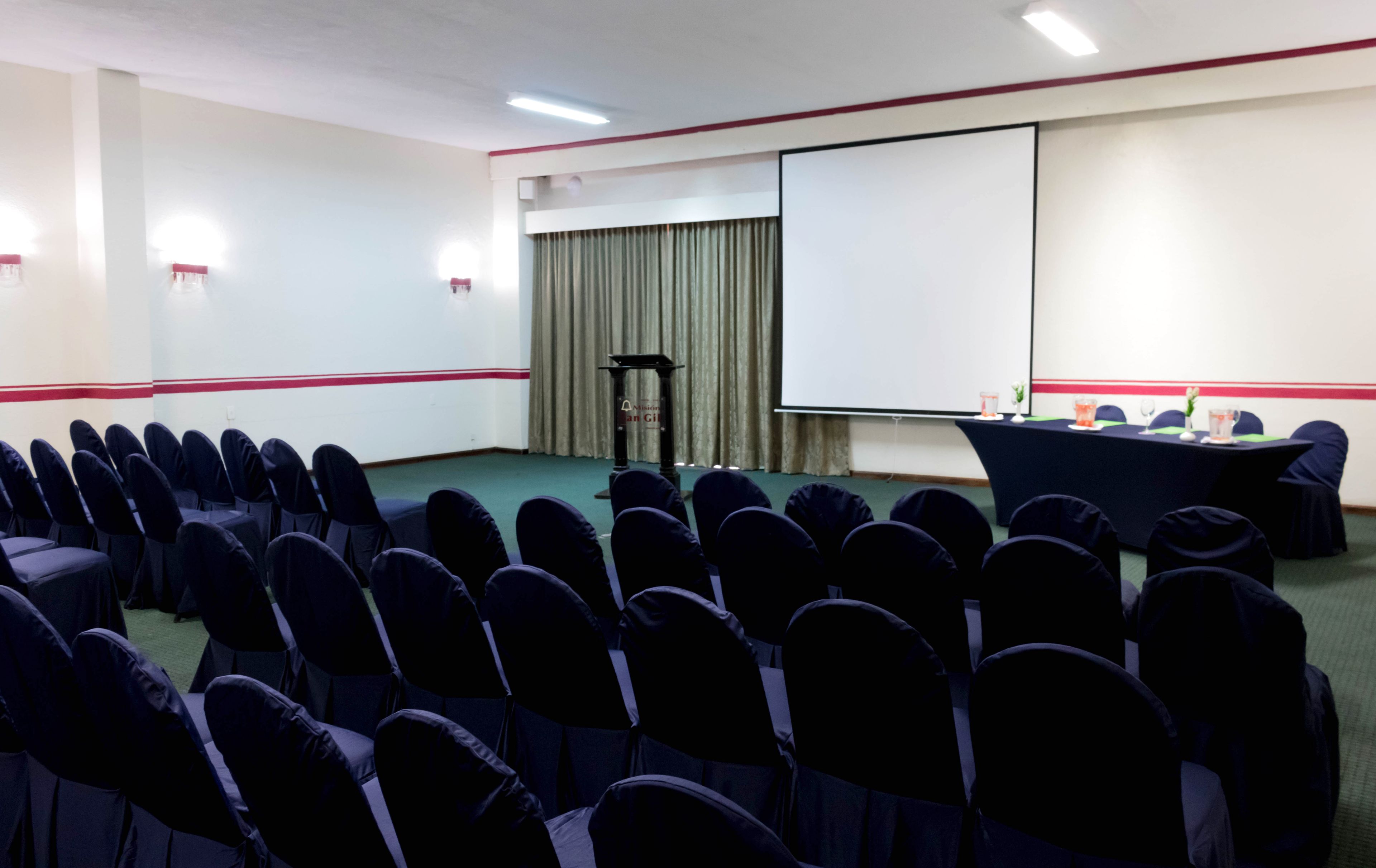 Un salón de conferencias con filas de sillas, un atril, una pantalla de proyección y una mesa con bebidas.