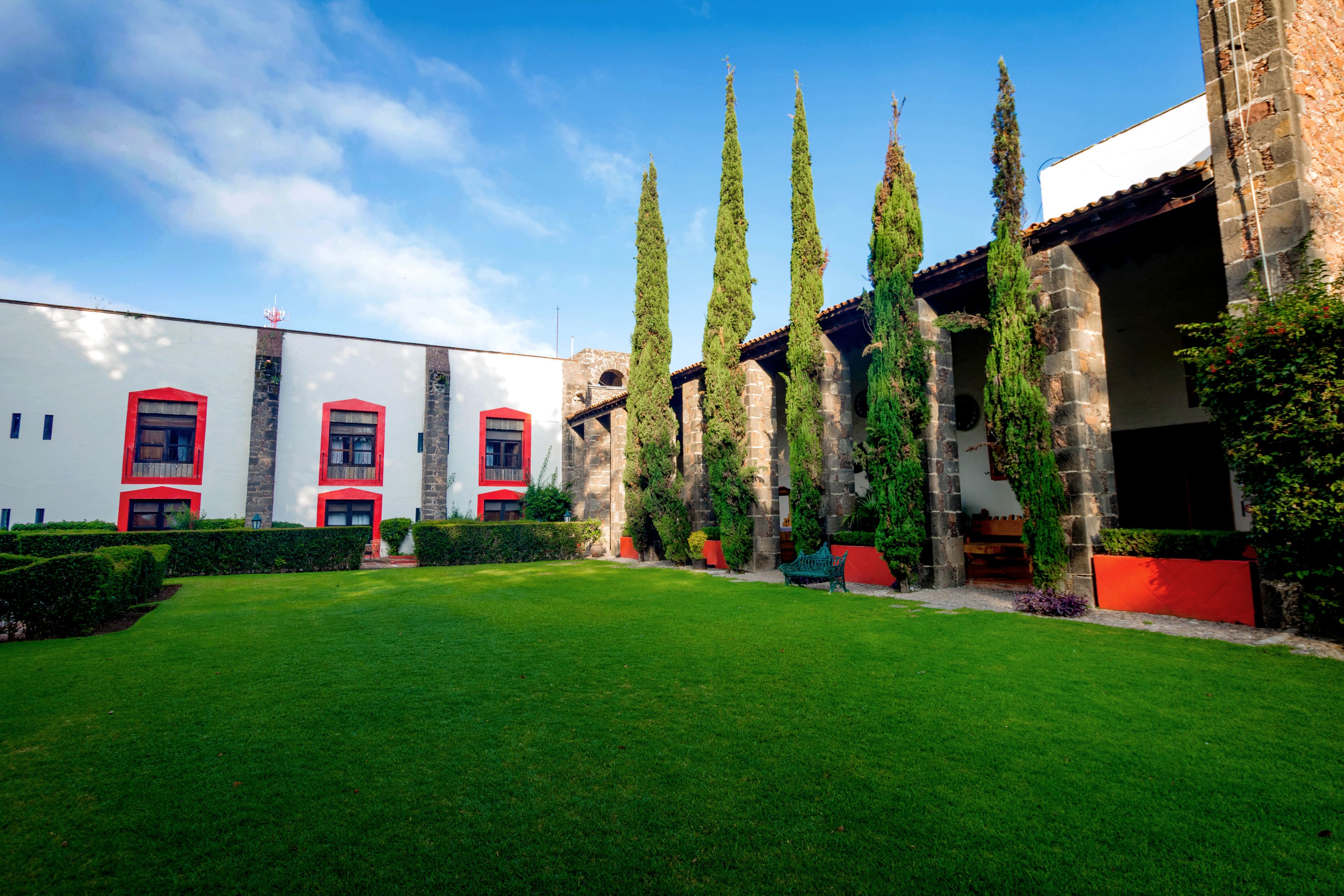 A courtyard with a white building featuring red accents, surrounded by well-maintained hedges and tall cypress trees.