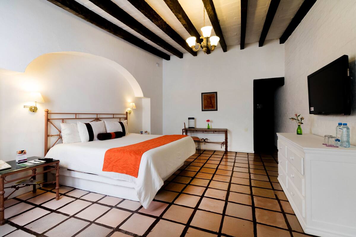 Habitación de hotel con cama, mesita de noche, lámpara, televisor, cómoda y suelo de baldosas.