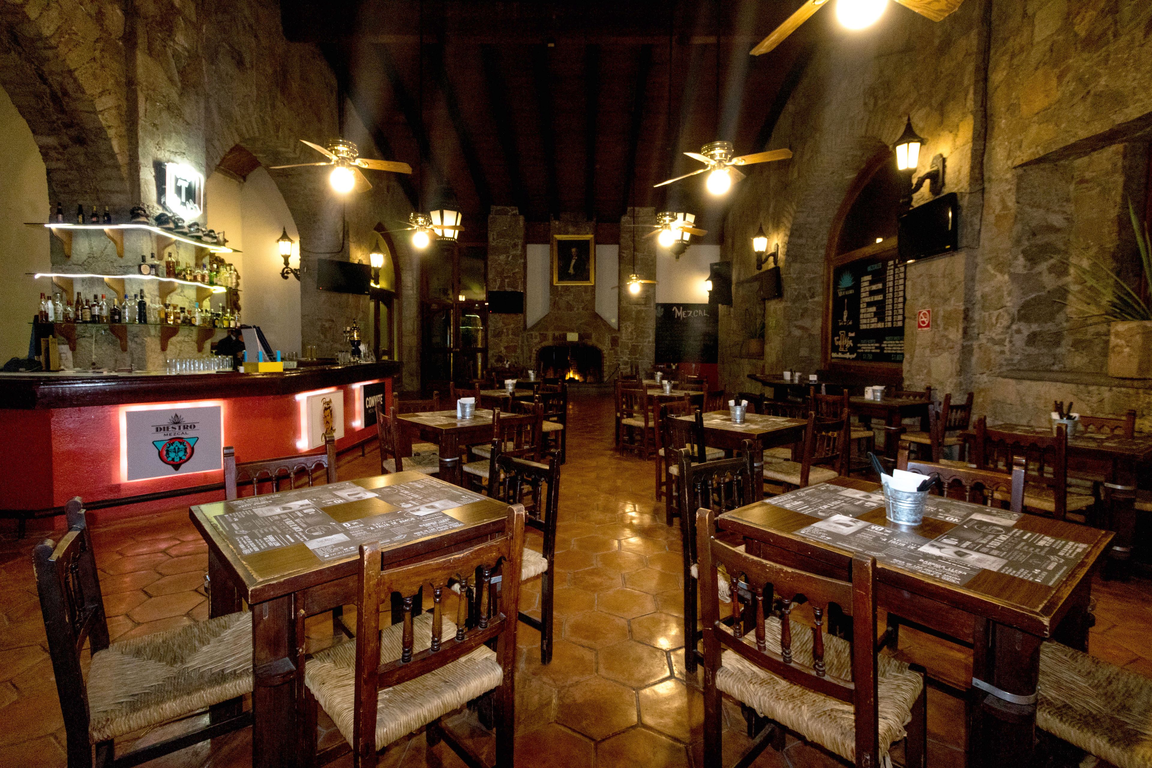 A restaurant with wooden tables and chairs, a bar area with shelves of bottles, and a stone wall.