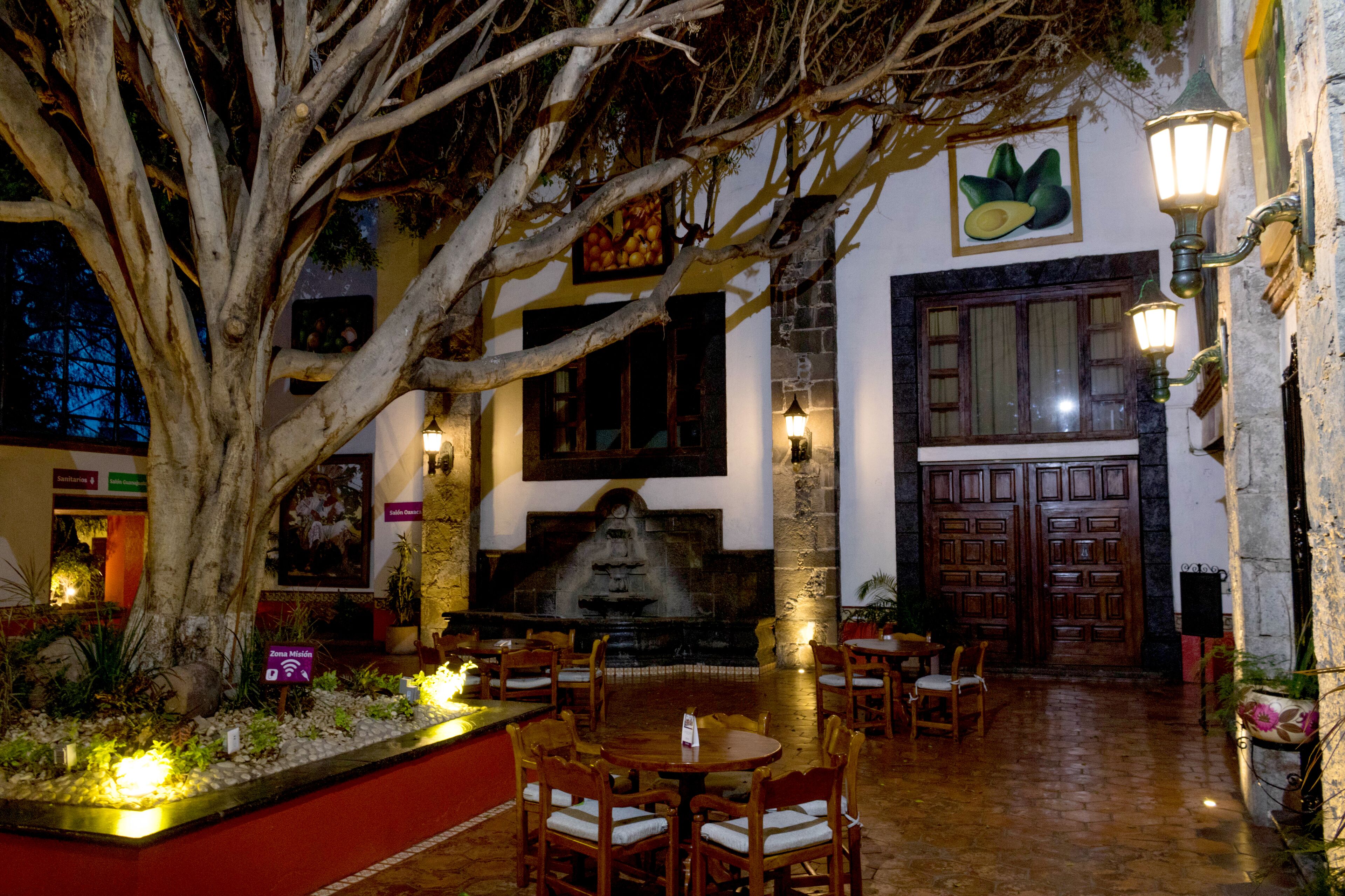 A courtyard with a large tree, stone fountain, wooden tables and chairs, and a stone wall with a door.
